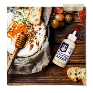 Double-strength liquid microbial rennet bottle beside a baked wheel of soft cheese topped with honey and chopped nuts, served with bread and grapes.