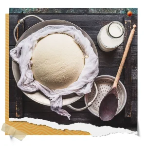 Fresh cheese curd draining in cheesecloth over a metal bowl beside a bottle of milk, wooden spoon, and strainer.