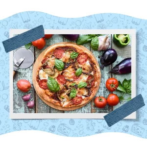 Homemade vegetable pizza topped with mozzarella, tomatoes, eggplant, basil, and peppers, surrounded by fresh vegetables and herbs.