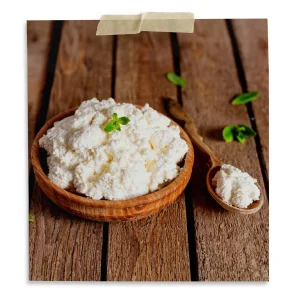 Bowl of fresh homemade cottage cheese on a rustic wooden table with a wooden spoon and mint garnish.